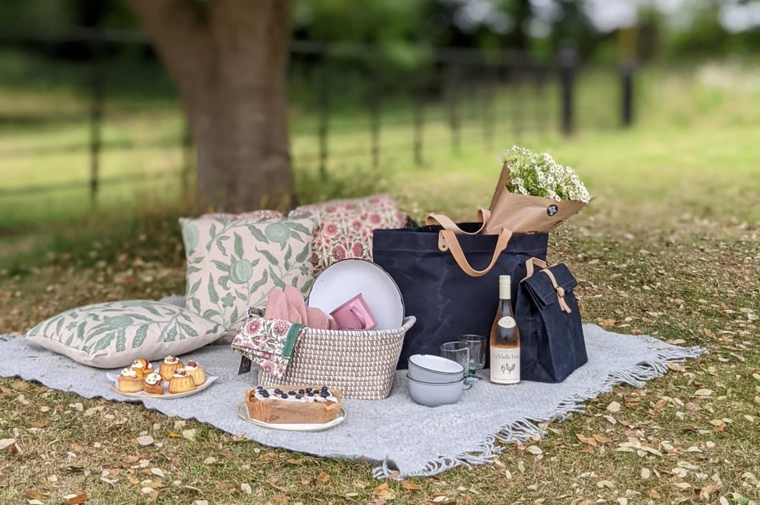 A range of Pod & Pip products laid out on a picnic blanket in a park, including Pod & Pip cushions, picnic bags, rugs, tinware and Alix Fauvel’s cakes.